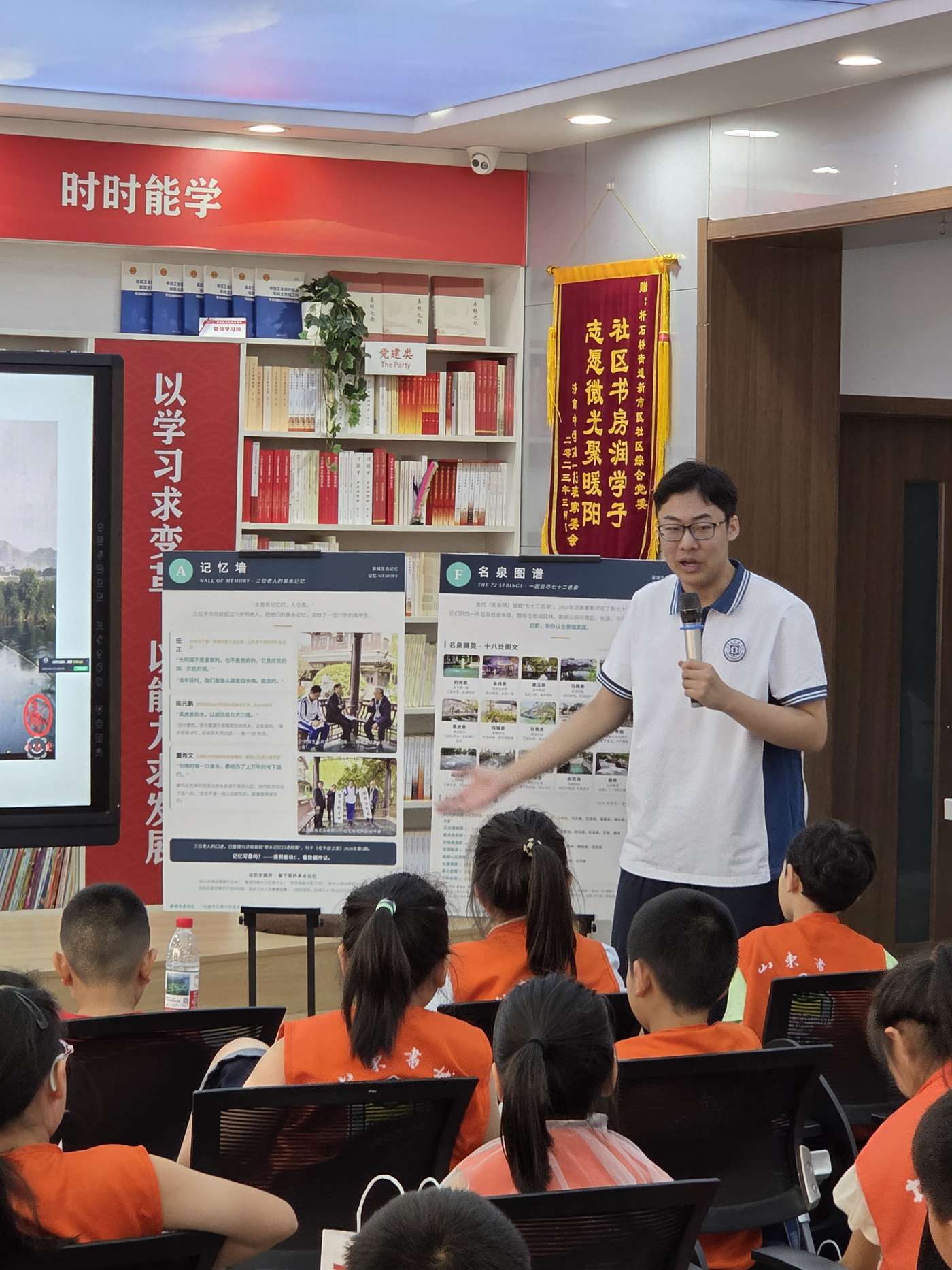 Ronghao Yang guiding young visitors through the exhibition panels, with a commemorative banner from the community committee on the wall