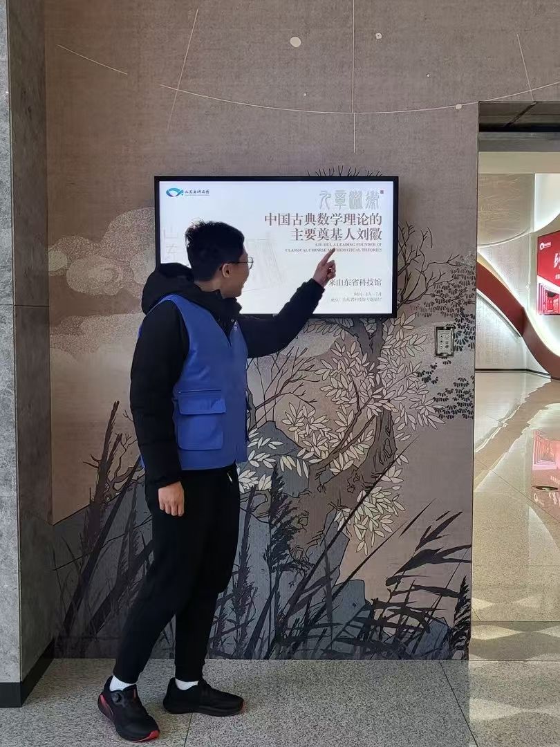 Volunteer docent in a blue vest presenting the Liu Hui exhibit screen at Shandong Science and Technology Museum