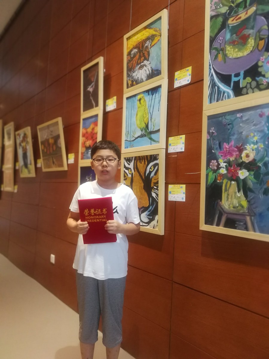 Young Ronghao holding an honorary certificate in front of a gallery wall hung with framed children's paintings, including his own