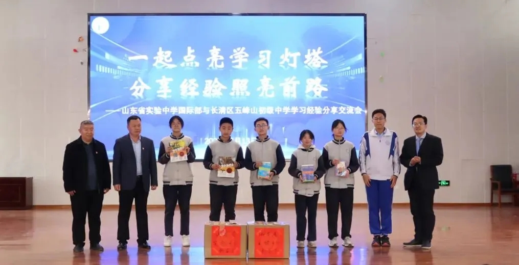 Pupils of Wufengshan Junior High School holding donated books in front of a screen reading “Lighting the Study Beacon Together”, with two cartons of donated books on the stage