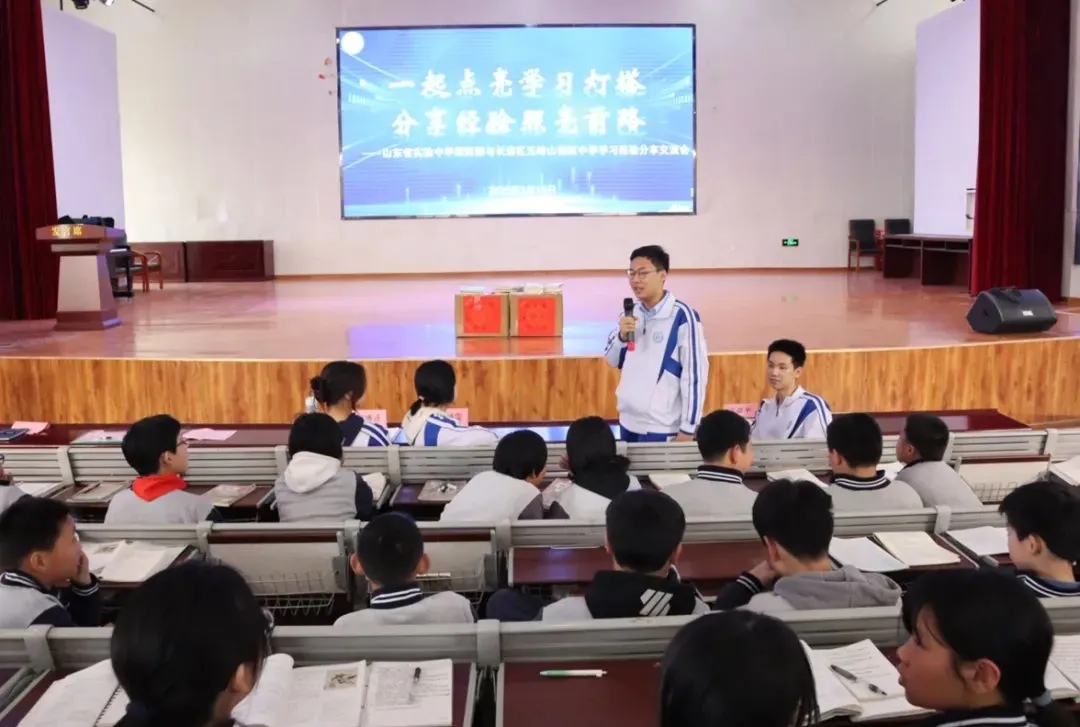 Ronghao Yang speaking into a microphone from the stage while Wufengshan pupils listen with notebooks open